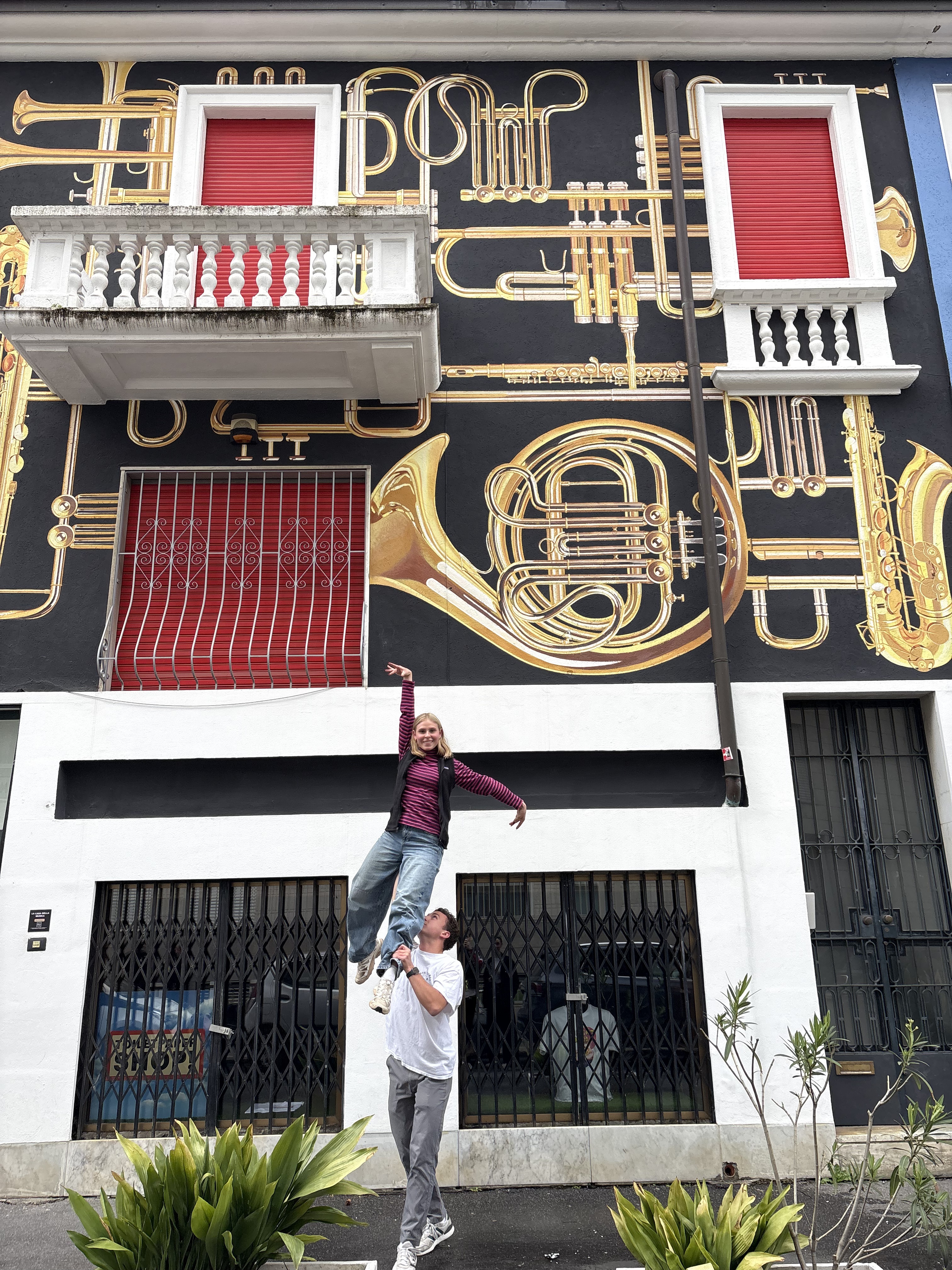 Male student lifting female student in a ballroom style lift standing in front of a building with musical instruments painted on the front.